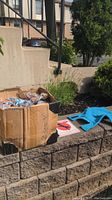 Box containing multiple rope chew toys, a dog training book, and a blue dog jacket placed on a stone ledge outdoors near some plants and stairs.