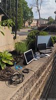 Photo showing multiple electronic devices including monitors and a tablet along with assorted cables placed on a stone ledge outdoors.