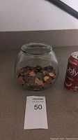 Glass fish bowl partially filled with pennies on a countertop next to a drink can for scale