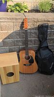 Photo of Denver acoustic guitar leaning against a stone wall with its black soft case beside it and the wooden percussion box on the left.