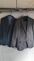 Two men's suit jackets hanging on hangers, one dark gray with patterned lining and one light gray with plain lining
