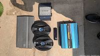 Top view of two car audio amplifiers, two car speakers, a small electronic device, and a blue power inverter on concrete