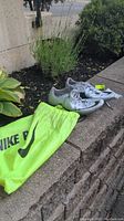 Pair of white Nike track spike shoes and neon yellow Nike drawstring bag displayed outdoors on a concrete ledge with plants in the background.