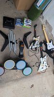 View showing PlayStation 4 console without cables, stack of video games, three guitar controllers and Rock Band drum set with stands and foot pedal on floor.