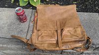 Front view of light brown leather Roots backpack with flap, two front pockets, shoulder strap, and visible wear, with a can for scale.