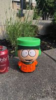 Front view of the Kyle Broflovski piggy bank placed outdoors next to a soda can for size reference. The figure features bright green hat and orange jacket.