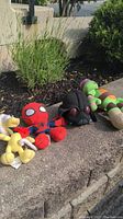 Photo shows four stuffed toys outdoors on a cement edge near plants: Spider-Man, black creature, green-orange turtle, and yellow character, all in a row.