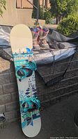 Salomon Lotus 146cm snowboard with teal Ride bindings upright against a stone wall, Thirty-Two women's snowboard boots size 8 visible in background along with a Burton bag and gloves.