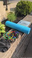 Photo showing blue yoga mat rolled up next to multicolored resistance bands on a concrete ledge outdoors.
