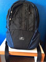 Front view of the Spire backpack showing black fabric with navy blue side panels and front zipper compartments.