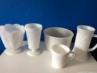 Five white milk glass items arranged against a blue background including scalloped bowl, two grape pattern goblets, small cup, and tall handled pitcher.