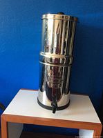 Front full view of polished stainless steel Berkey water filtration system with black spigot on white table against blue wall.