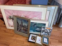Photo showing a group of framed prints and paintings leaning against a couch with a wooden floor; includes a large pink floral print, a scenic town painting, nature scene painting, small black and white framed artwork, and three small framed prints in foreground.
