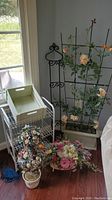 Photo showing metal trellis with peach-colored faux flowers, wire shelf with wooden box tray on top, and three faux flower arrangements on floor in front.