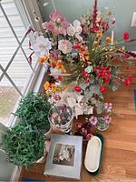 Photo showing multiple colorful faux flower arrangements in glass and ceramic vases along with additional decorative vases and plants on hardwood floor near window