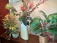 Overview of three faux flower arrangements on a wooden surface including vases with various artificial flowers and plants.
