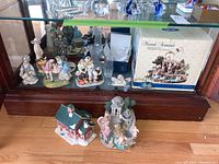Wide shot of ceramic figurines including Norman Rockwell baseball figurines, musical angel and cherubs sculpture in original box, and various other figurines in glass cabinet.