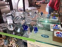 Display of clear and blue glass figurines and paperweights on glass shelves including birds, fish, elephant and boxed octopus pieces.