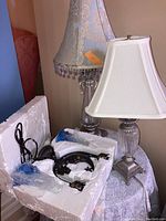 Two different table lamps displayed together with cords and packaging visible. One lamp has a white rectangular shade and clear glass base; the other has tall blue floral shade with bead trim and ornate base.