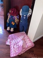 Photo showing two rag doll figurines standing against a wall next to the folded pink and white quilt on the floor