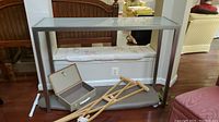 Sofa table with frosted glass top and metal base shown from front angle with surrounding items including crutches and a small open box.