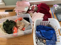 Large clear tub containing folded fabric pieces in green, pink, beige, black colors including seat covers and chair covers, placed next to plastic laundry baskets with additional fabrics and linens.