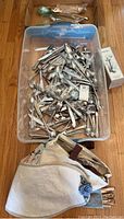 Plastic container with assorted metal cutlery including spoons, forks, knives, along with fabric pouch potentially for utensil storage. Items show signs of tarnish and require cleaning.