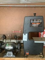 Black & Decker band saw and Ohio Forge bench grinder placed side by side on a shelf with pegboard behind them.