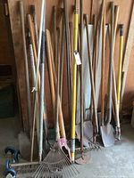 Photo showing a variety of garden tools leaning against a wall including rakes, shovels, hoes, a pitchfork, and four caster wheels on the floor.