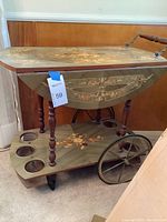 Side view of vintage bar cart featuring floral painted top, drop-leaf, turned legs, and bottle holders on lower shelf with mixed wheels for mobility.
