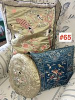 Large rectangular pillow with embroidered peacock and birds among flora. Pale gold color with patterned silk border. Positioned on a patterned couch.