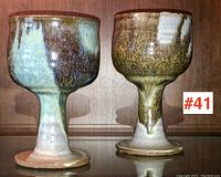 Front view of two pottery goblets standing side by side on a reflective surface with a wooden background. Shows overall shape and glaze details.