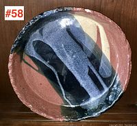 Top-down photo of circular pottery plate showing abstract glaze design in black, gray, and cream on red-brown base.