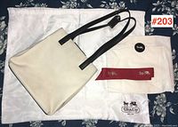 Image showing the white Coach handbag with black leather handles, placed on a floral fabric background with original Coach dust bag, red ribbon, and a round Coach branded item.