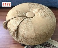 Side angle of round ottoman with tassel ends resting on wooden floor and patterned area rug.