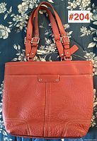 Front view of vintage Coach leather handbag on floral background, showing embossed Coach logo patch and double buckle straps.