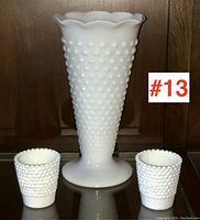 Front view of the vintage hobnail milk glass tall vase flanked by two small hobnail milk glass cups on a reflective surface with wooden background.
