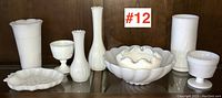 Set of 9 white vintage milk glass pieces arranged on a wooden shelf: vases, bowls, goblets, and an oval dish with scalloped edges.