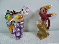 Two colorful vintage ceramic bird items, wall pocket on left with purple grapes, and vase on right with red bird and yellow wings.