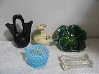 Five vintage items on a table: blue hobnail glass bowl with handle, black double spout green leaf-shaped double wedding vase, small pale yellow Zsolnay vase with floral design, two clear glass knife rests with textured pattern