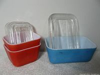 Three vintage Pyrex fridge containers with clear glass lids, two square red-orange and one rectangular sky blue, showing wear.