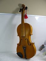 Full front view of the violin showing the spruce top with F-holes, fine wood grain, black fingerboard and pegs, and a red tassel on scroll.