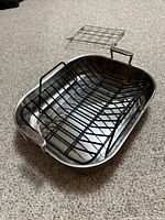 Stainless steel roasting pan with black metal roasting rack and additional smaller metal rack inside, shown from an angle on carpeted floor.