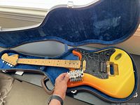 Electric guitar with sunburst body and maple neck lying inside blue plush-lined hard case, strings loose, one pickup missing.
