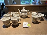 Overall view of one Brambley Hedge teapot surrounded by eight matching teacups on countertop