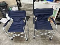 Front view of two blue foldable camping chairs side by side showing fabric seats, backs, and armrests on metal frames.