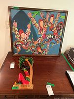 Framed abstract painting on canvas with figures with raised hands and wooden Jamaica wall hanging on table
