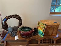 Wide view of lot atop wooden furniture including the stone elephant sculpture, ceramic bowl, horse bookend, bright modern painting, and woven basket box.
