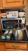 Photo of the tea kettle, refrigerator water filters, and assorted mugs on kitchen counter in front of microwave. Items show varying degrees of use and packaging intact.