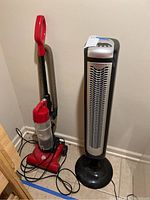 Photo showing the Dirt Devil vacuum cleaner in red with hose and power cord alongside the Bionaire tower fan in black and silver, standing on tiled floor.